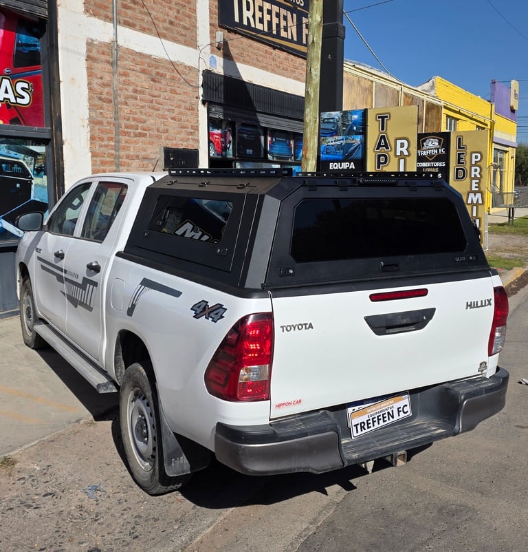 White Toyota Hilux pickup truck with black canopy parked on street with brick buildings in background
