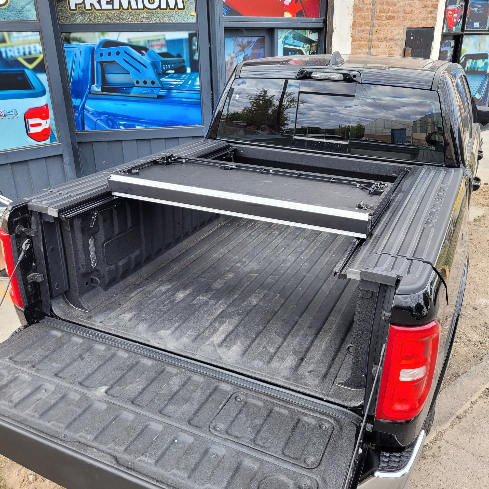 Black pickup truck with open bed displaying tonneau cover and aluminum crossbars in front of storefront