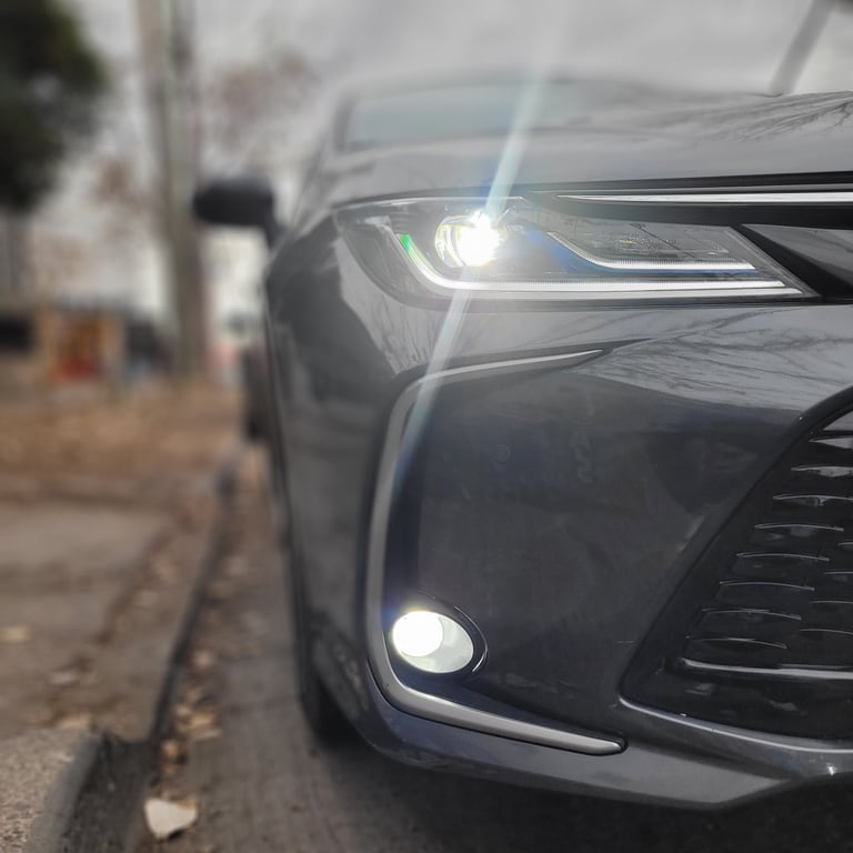 Close-up of a dark gray cars front end showing LED headlights and fog lights illuminated on a street