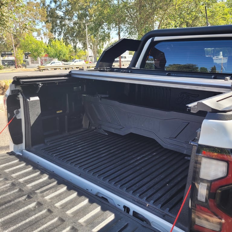 Open pickup truck bed with black tonneau cover partially rolled back, showing ridged cargo area and tailgate on a sunny suburban street