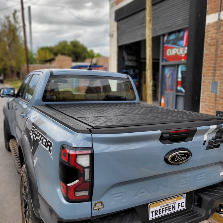 A silver Ford Ranger pickup truck with tonneau cover parked outside a brick garage, with custom license plate and Raptor badging visible.