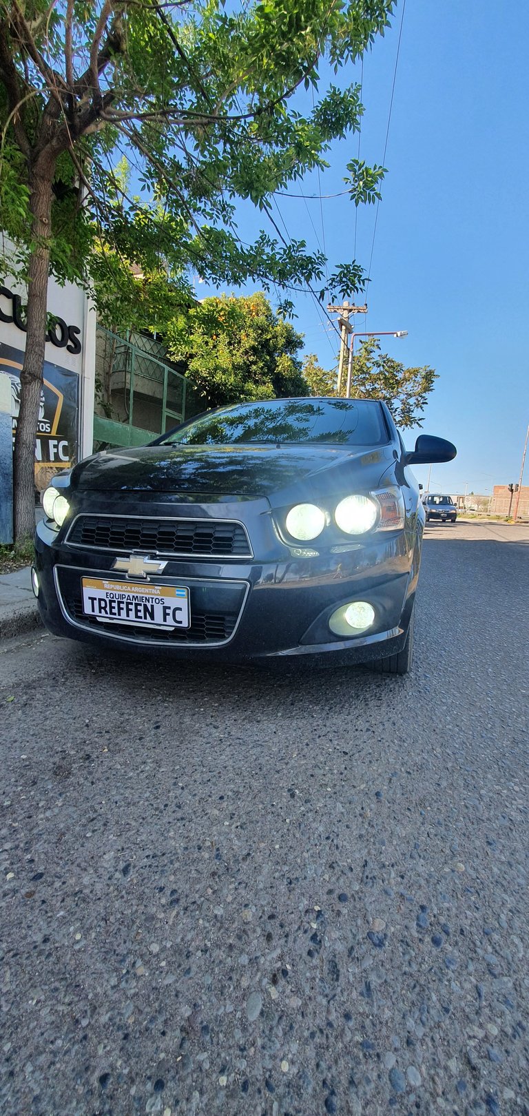 Dark gray Chevrolet Sonic with Treffen FC license plate parked on street under tree with clear sky