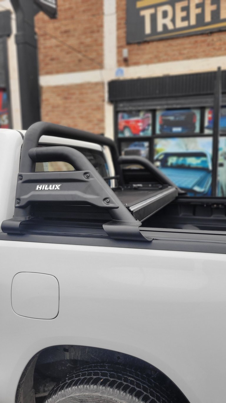 Close-up of a black Hilux tonneau cover on a white pickup truck bed parked in front of a storefront
