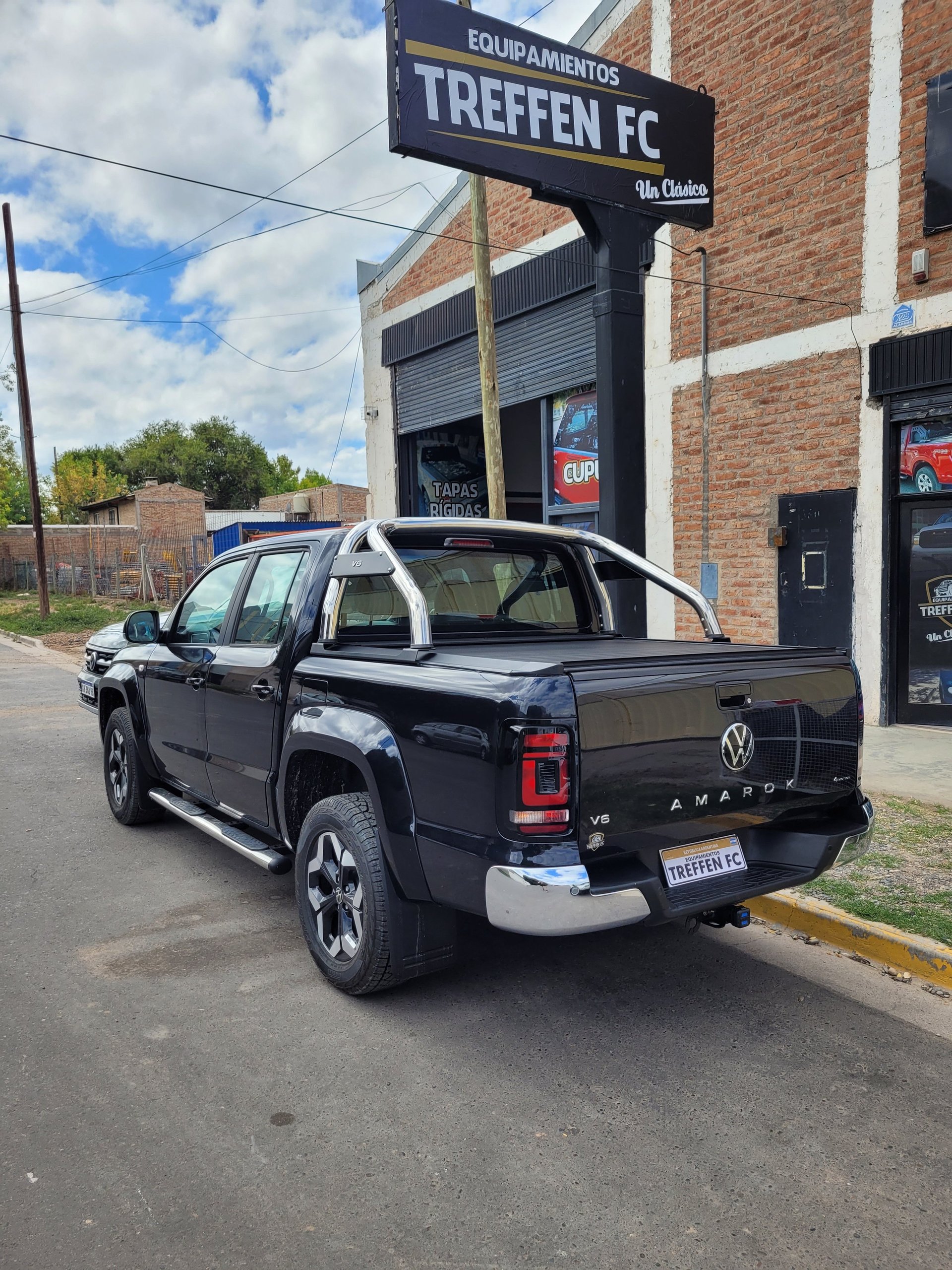 Black Volkswagen Amarok pickup truck parked on street in front of a brick building and Treffen FC equipment shop sign