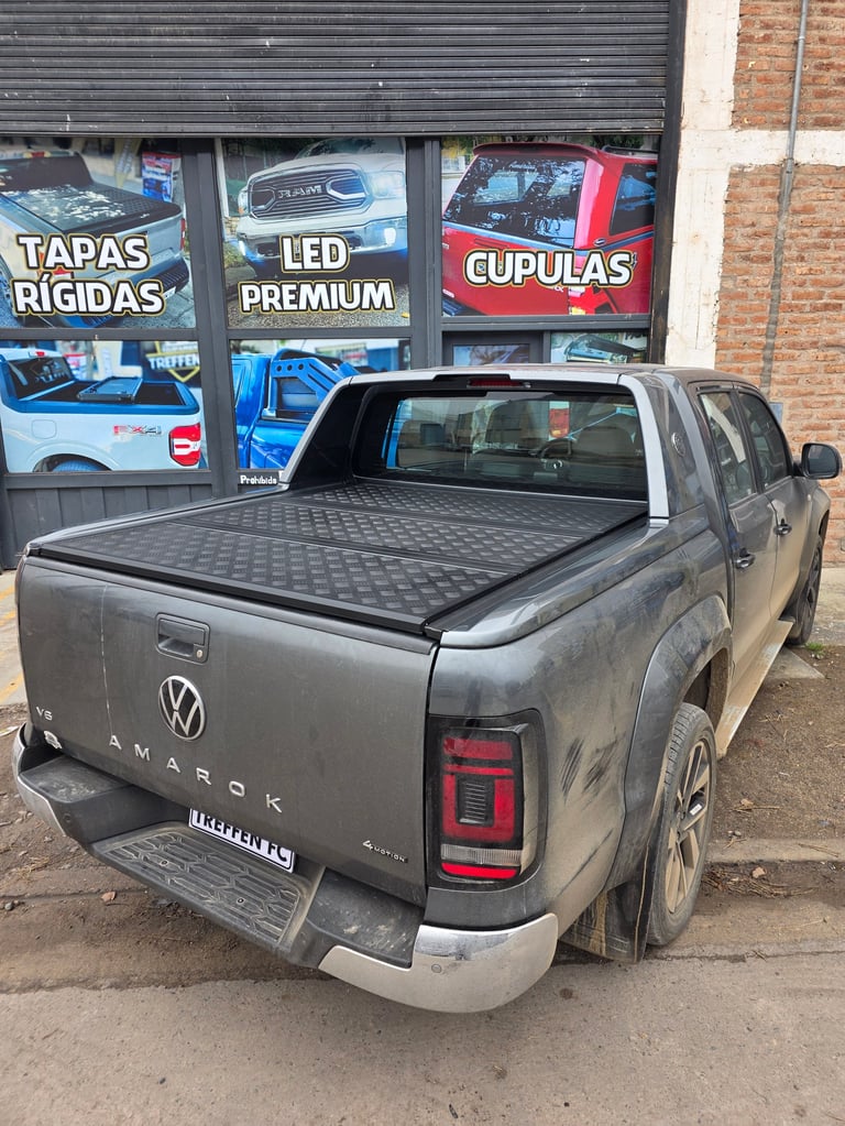 Gray Volkswagen Amarok pickup truck with black tonneau bed cover parked in front of auto parts shop with Spanish signage