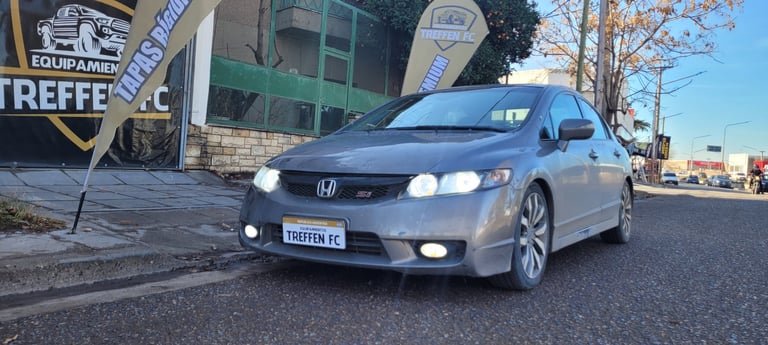 Silver Honda Civic coupe parked on street with headlights on, near commercial signage and buildings