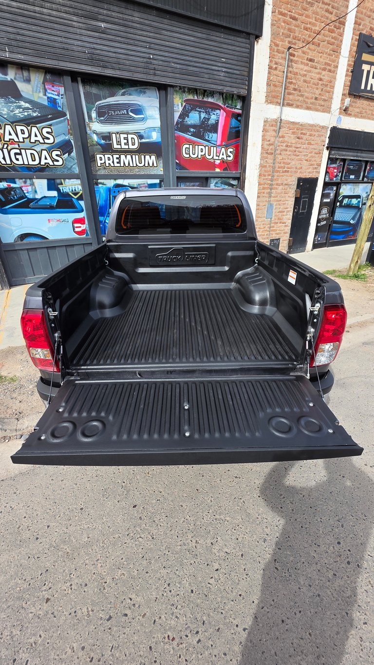 Black pickup truck with open tailgate showing rubber bed liner and retractable tonneau cover, parked in front of auto accessories shop