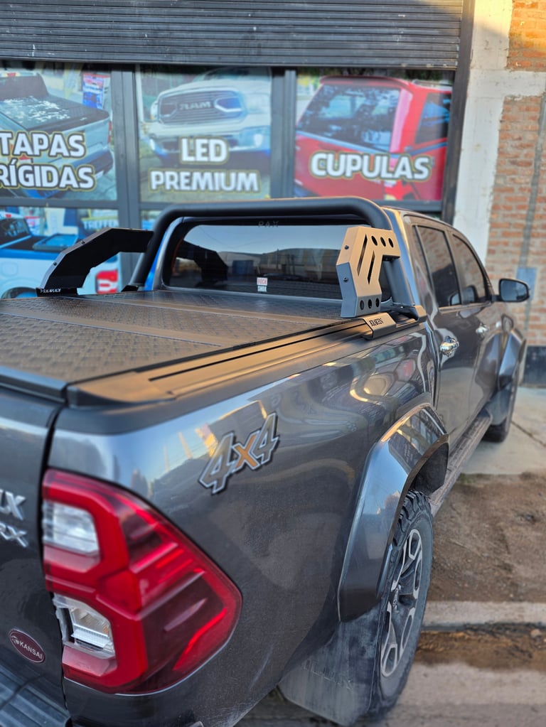 Dark pickup truck with open tailgate and tonneau cover at a storefront with signs for tapas, LED lights, and car accessories