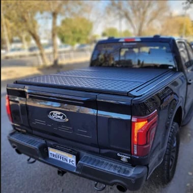 Black Ford pickup truck with blue tonneau cover on truck bed, parked in driveway with trees in background