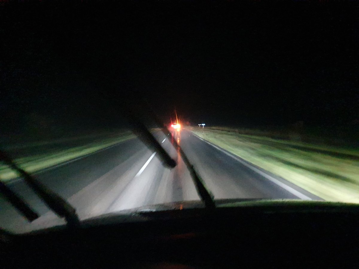 View from a moving vehicle at night following a car with bright headlights on a straight highway