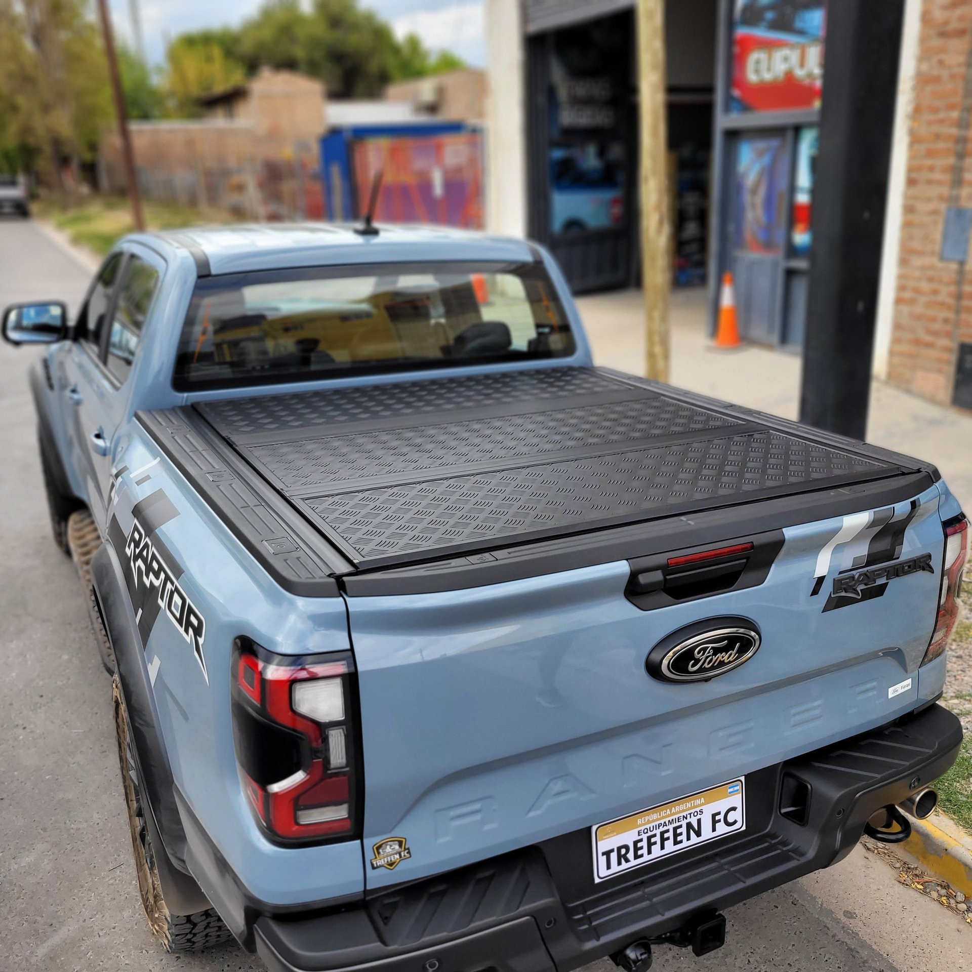 Light blue Ford Ranger pickup truck with black tonneau cover in a parking lot with commercial buildings