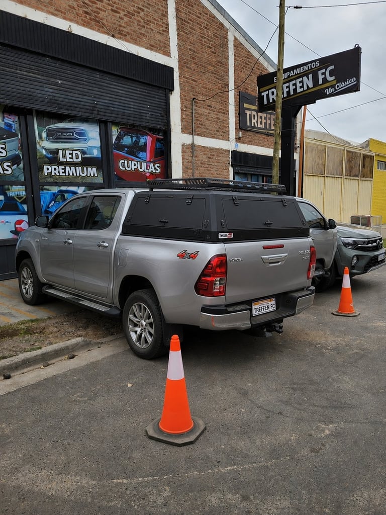 Silver pickup truck parked on street with orange traffic cones, brick building and Triffee FC sign visible in background