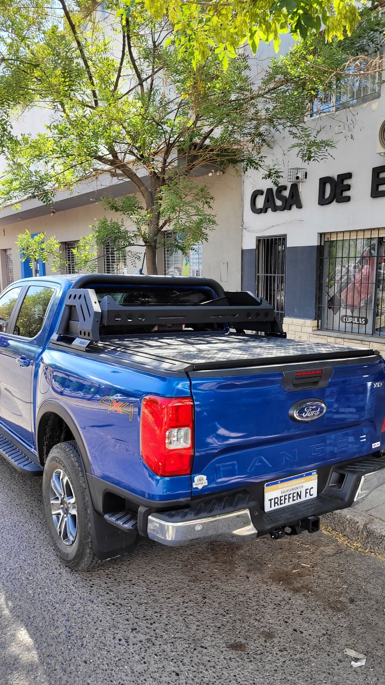 Blue Ford Ranger pickup truck with tonneau cover parked on a street near a white building with a tree overhead