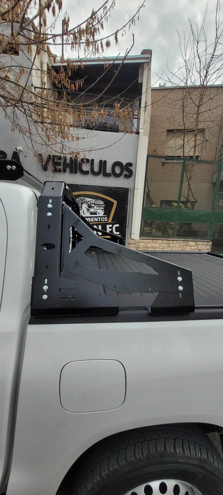 White pickup truck bed with black tonneau cover, parked near building with vehiculos sign and dried vines overhead